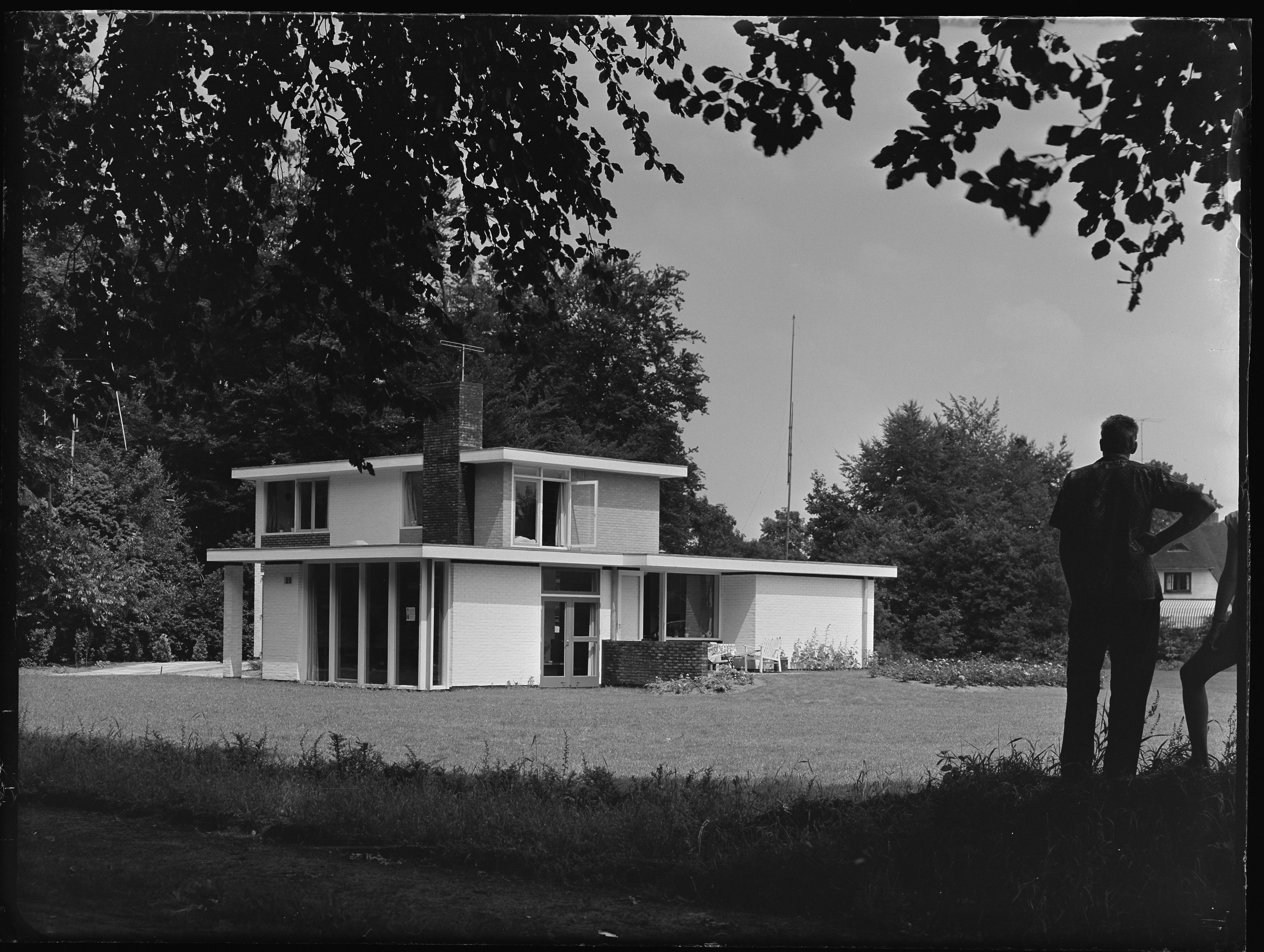 Vrijstaand huis van twee verdiepngen met plat dak, rechts staan twee mannen het huis te bekijken.